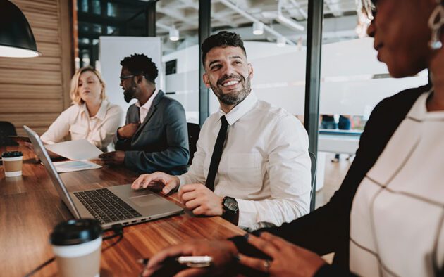 Four people in business attire having a discussion around a table with laptops and coffee cups.