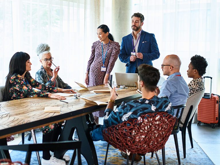 A group of people sitting and standing around a table in a meeting, engaging in discussion.