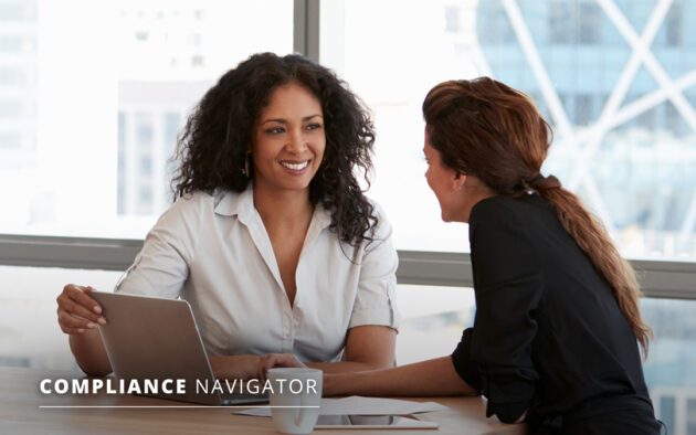 Two women in business clothing sitting at a table while working on a laptop
