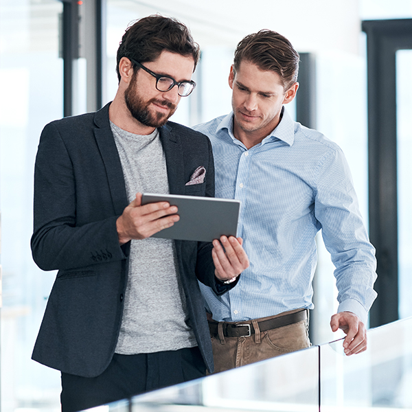 Two men standing in an office, looking at a tablet together. One is wearing glasses and a blazer; the other is in a light blue shirt.