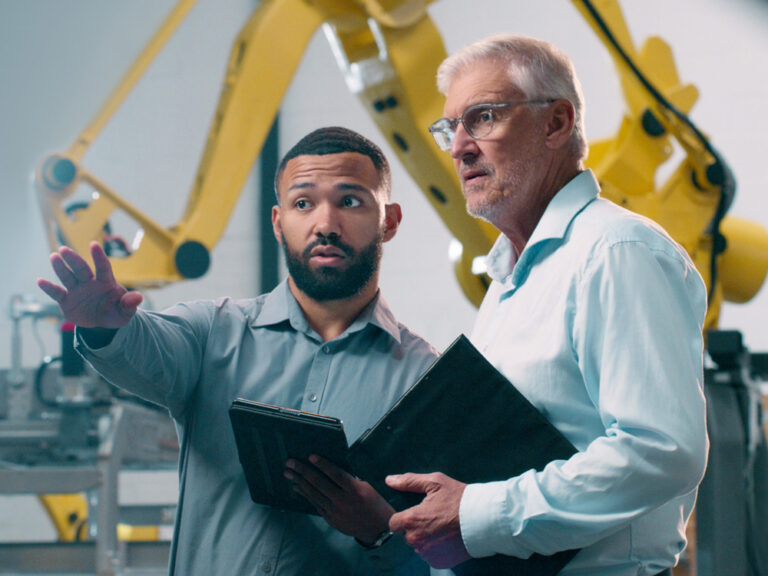 Two men in a factory setting, one gesturing while holding a tablet, the other holding a folder. Large yellow robotic arm machinery is visible in the background.