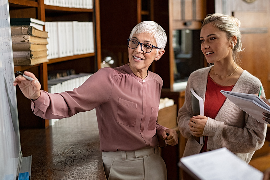Two women discussing in front of a whiteboard in an office, one pointing at the whiteboard while the other holds documents. Shelves with books are in the background.