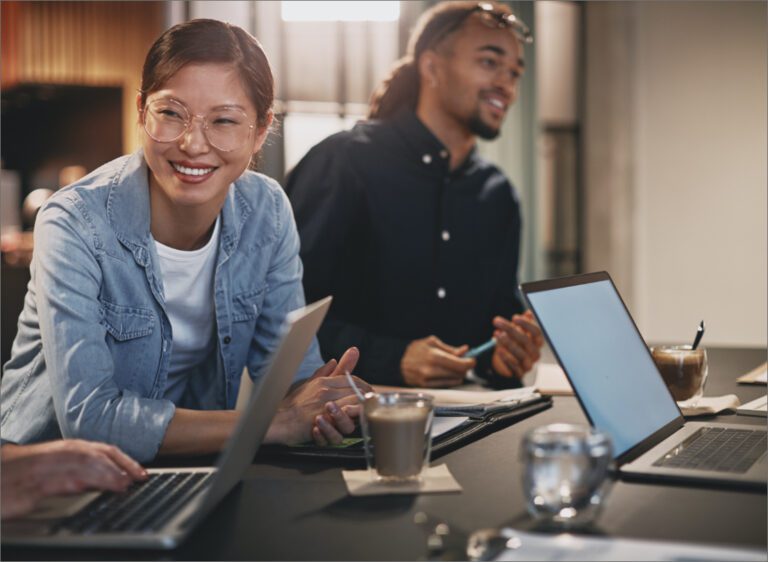 An Asian woman and a Black man are working with a team at a conference table, with several notepads and laptops nearby.