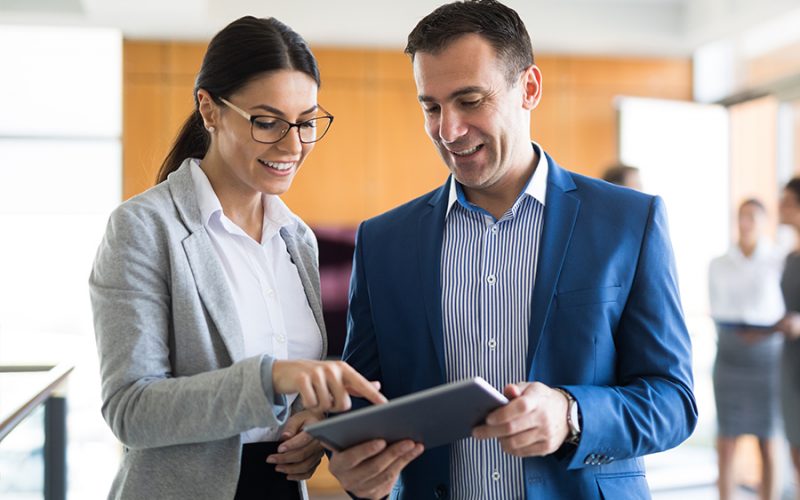 Two people, one in a gray blazer and the other in a blue suit, are standing and looking at a tablet together in an office setting.
