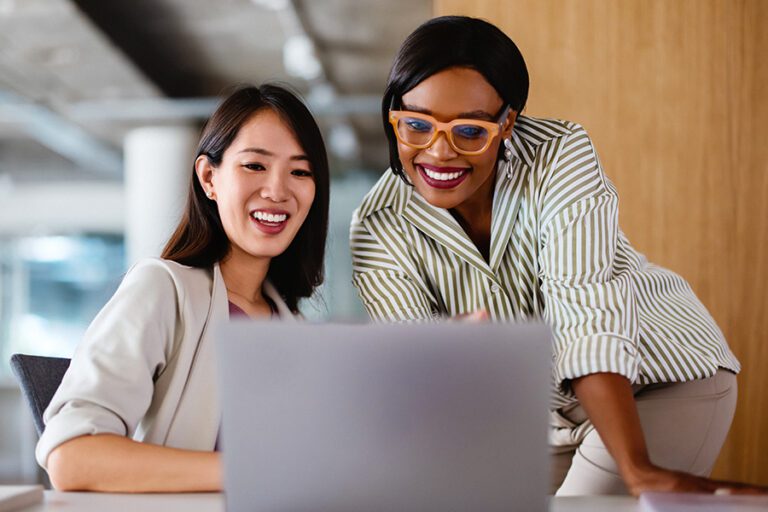 Two women in a modern office setting smile while looking at a laptop screen, one seated and one standing next to her.