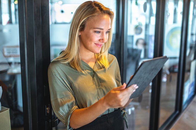 Woman in a green blouse stands in an office, looking at a tablet with a smile.