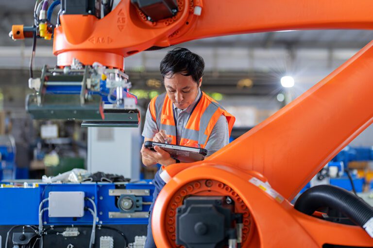 A worker in a safety vest uses a tablet near an industrial robot in a factory setting.