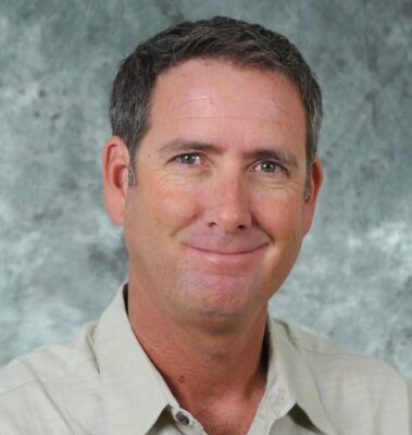 Man with short gray hair and a neutral expression, wearing a beige shirt. Neutral background.