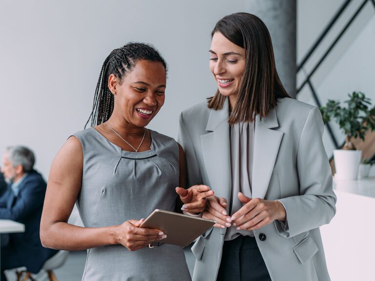 Two women in business attire smiling while looking at a tablet in an office setting.