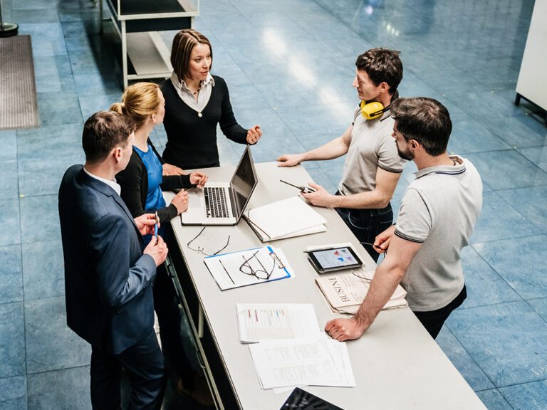 Six professionals in casual and semi-formal attire have a discussion around a long table with laptops, tablets, and documents in a modern office setting.