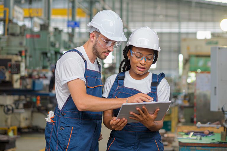 Two workers in hard hats and overalls discuss a project while looking at a tablet in an industrial setting.