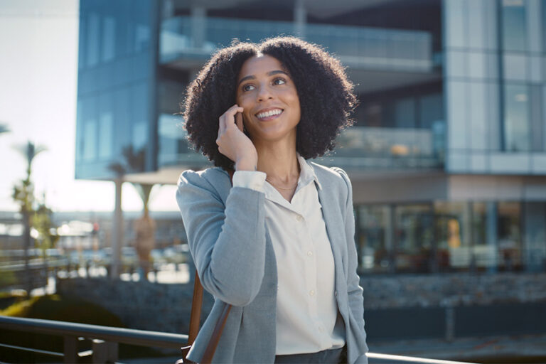 A woman in business attire smiles while talking on her phone outside a modern building.