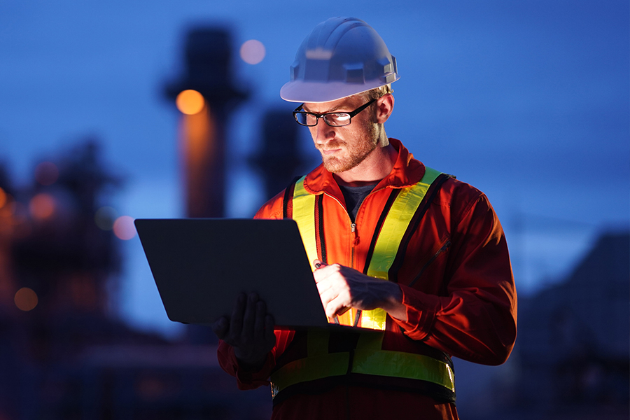 Engineer in a hard hat and high-visibility vest works on a laptop outdoors at dusk, with industrial structures in the background.