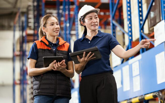 Two people are standing in a warehouse, one is in a hard hat and the other is in a vest.
