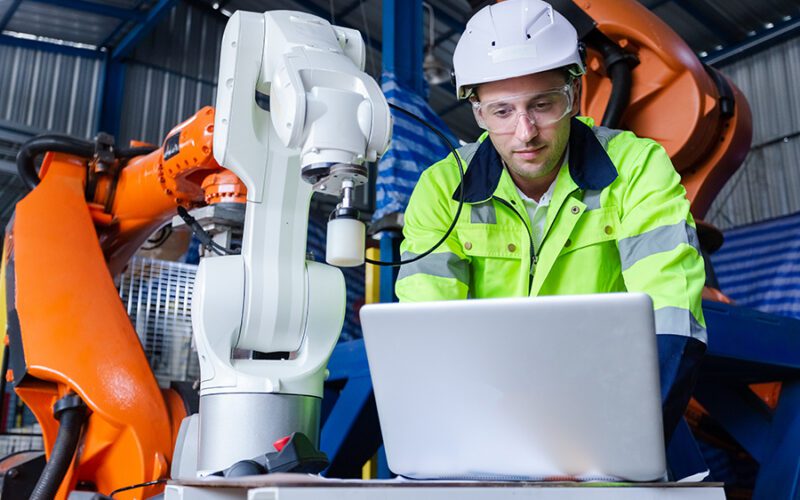 A worker in a high-visibility jacket and hard hat uses a laptop while standing next to industrial robots inside a factory setting.