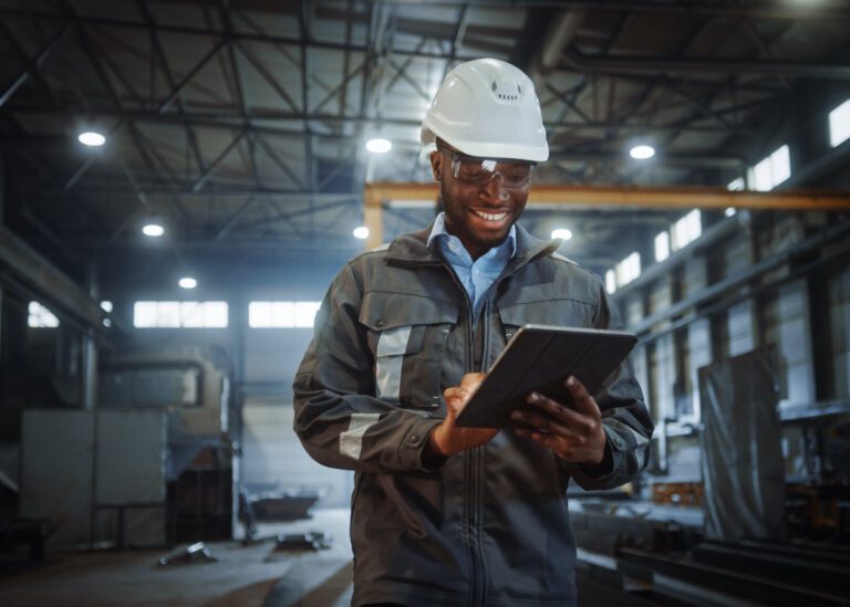 A warehouse worker with a hard hat on is smiling while typing on a tablet.