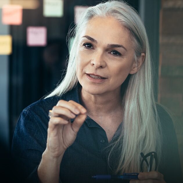 A woman with long gray hair speaks, holding a pen. She appears to be in a professional or educational setting, with sticky notes visible in the background.
